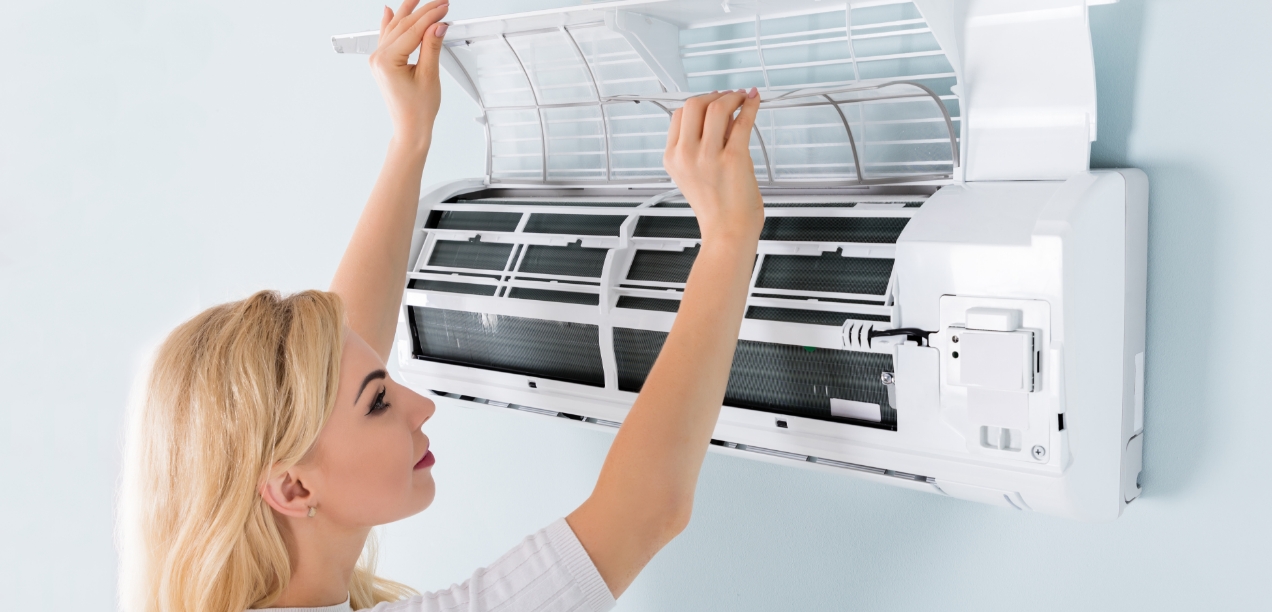 A young caucasian woman opens up a ductless heat pump indoor unit to check its filters.